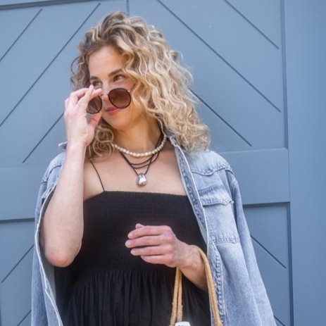 Woman wearing black tank top & denim jacket, standing in front of a blue door.